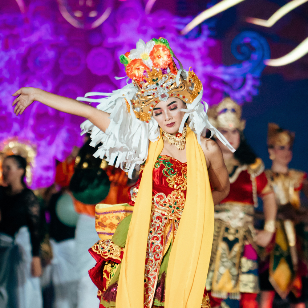 Deena wearing traditional Indonesian costume dancing on stage with her arm up on the air and her eyes closed